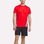 A man in a bright red Reebok Men's Train Blocked T-Shirt with white side accents and black shorts faces forward against a white background, highlighting stylish men’s activewear.