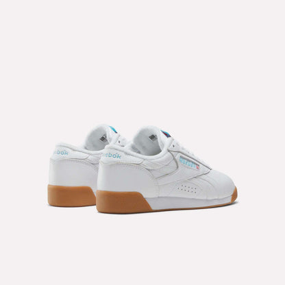 The Reebok Women's Freestyle Lo Shoes feature a white design with gum soles, light blue branding on the heel and side, and perforated details, shown against a plain white background.