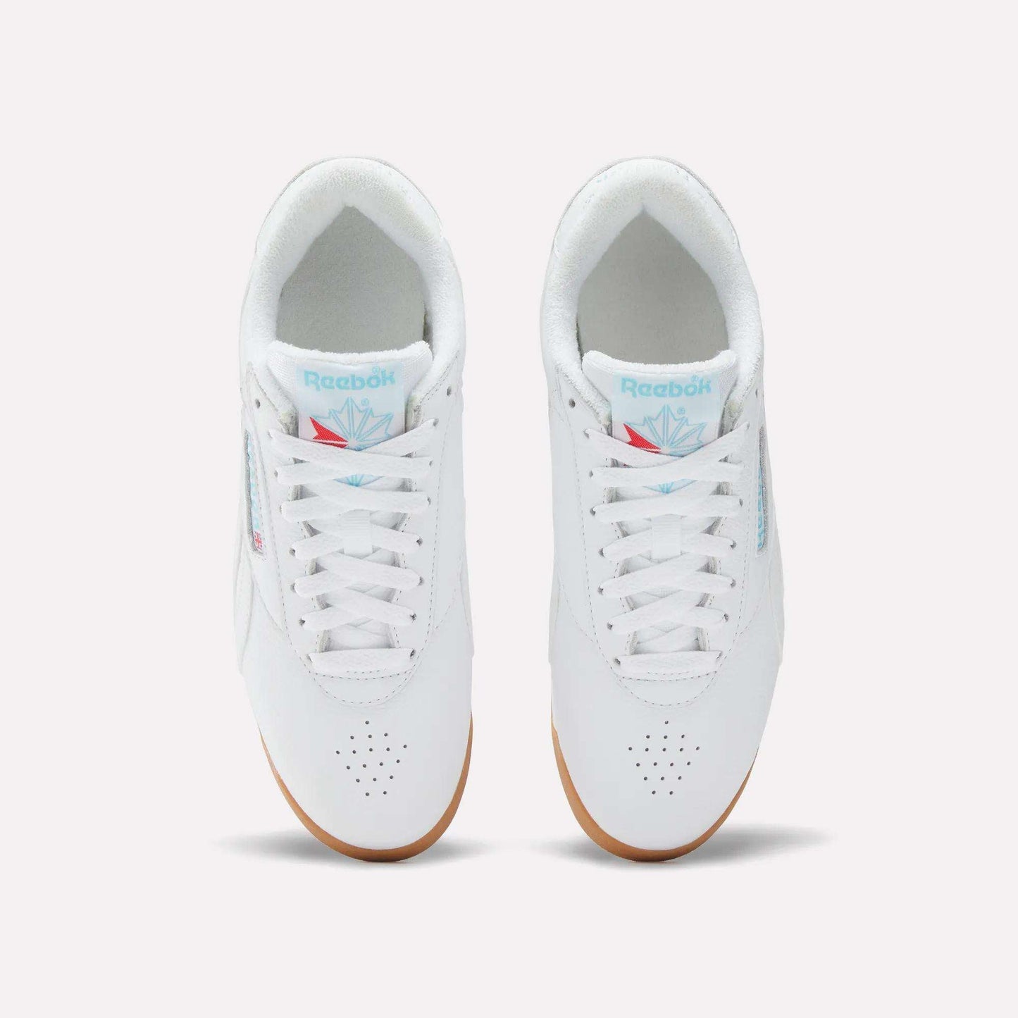 Top view of Reebok Women's Freestyle Lo Shoes in white with white laces, light blue branding on the tongue, perforated toe area, and light brown gum soles, set against a plain white background.