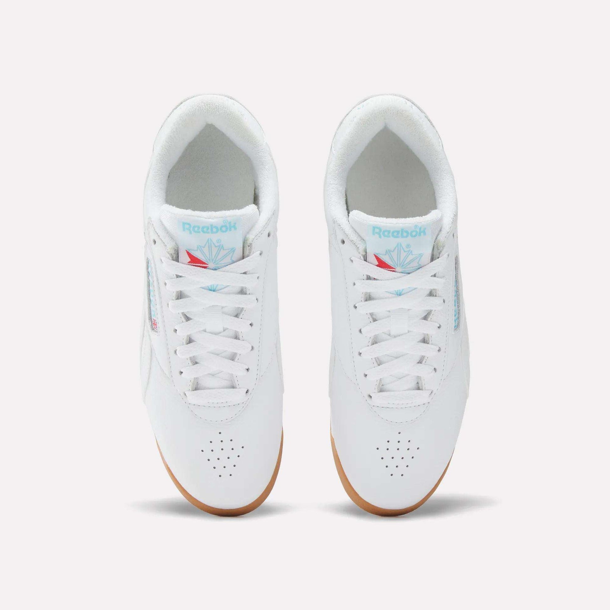 Top view of Reebok Women's Freestyle Lo Shoes in white with white laces, light blue branding on the tongue, perforated toe area, and light brown gum soles, set against a plain white background.