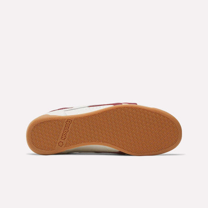 The image shows the bottom view of Reebok Women's Freestyle Lo Shoes with a gum rubber sole featuring a honeycomb pattern, the embossed Reebok logo near the heel, and a white leather upper with a touch of red.