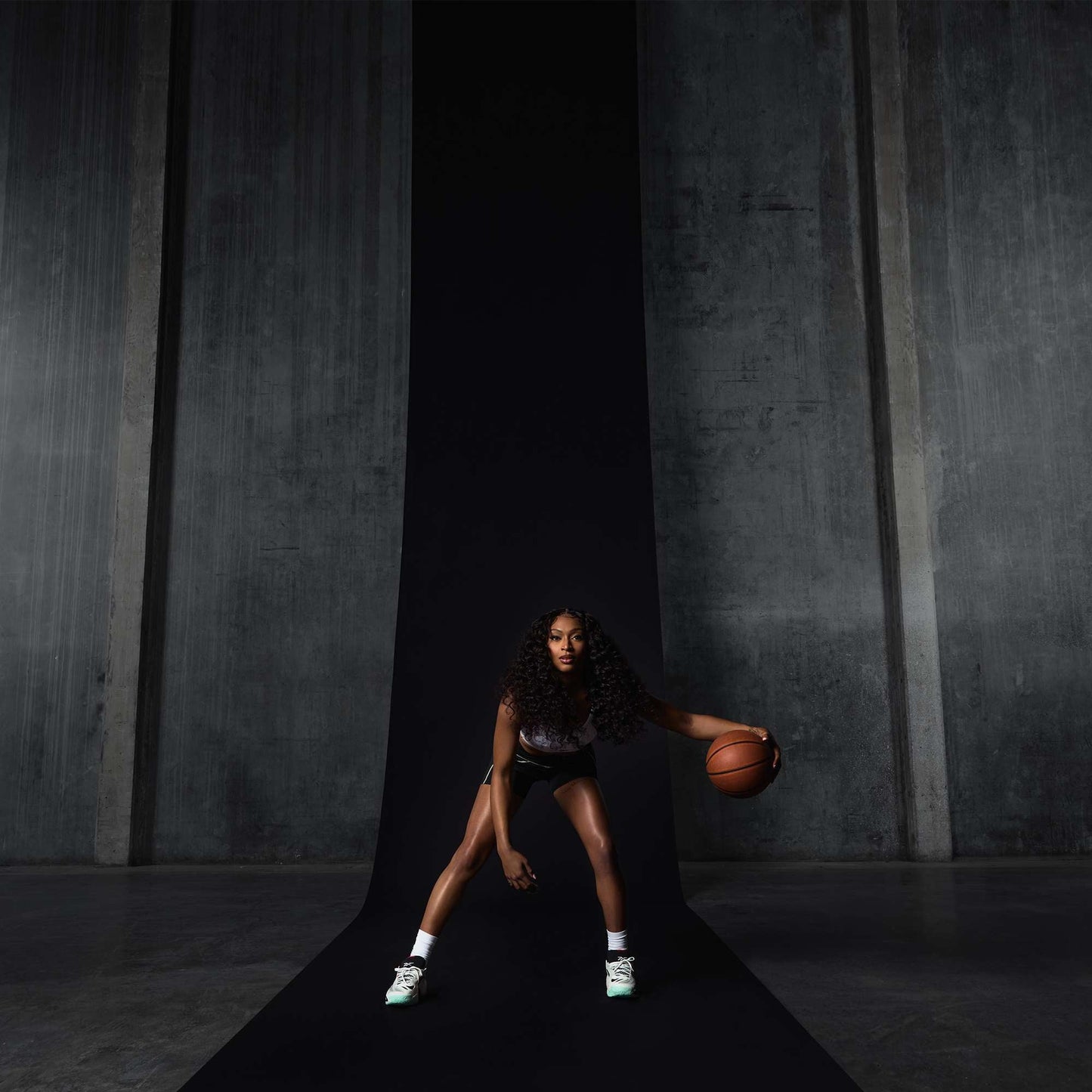 A woman with long curly hair dribbles a basketball on a black runway in front of gray concrete walls, wearing a sports outfit and Reebok Engine A 26 Basketball Shoes designed for energy return and stability, appearing focused and athletic.