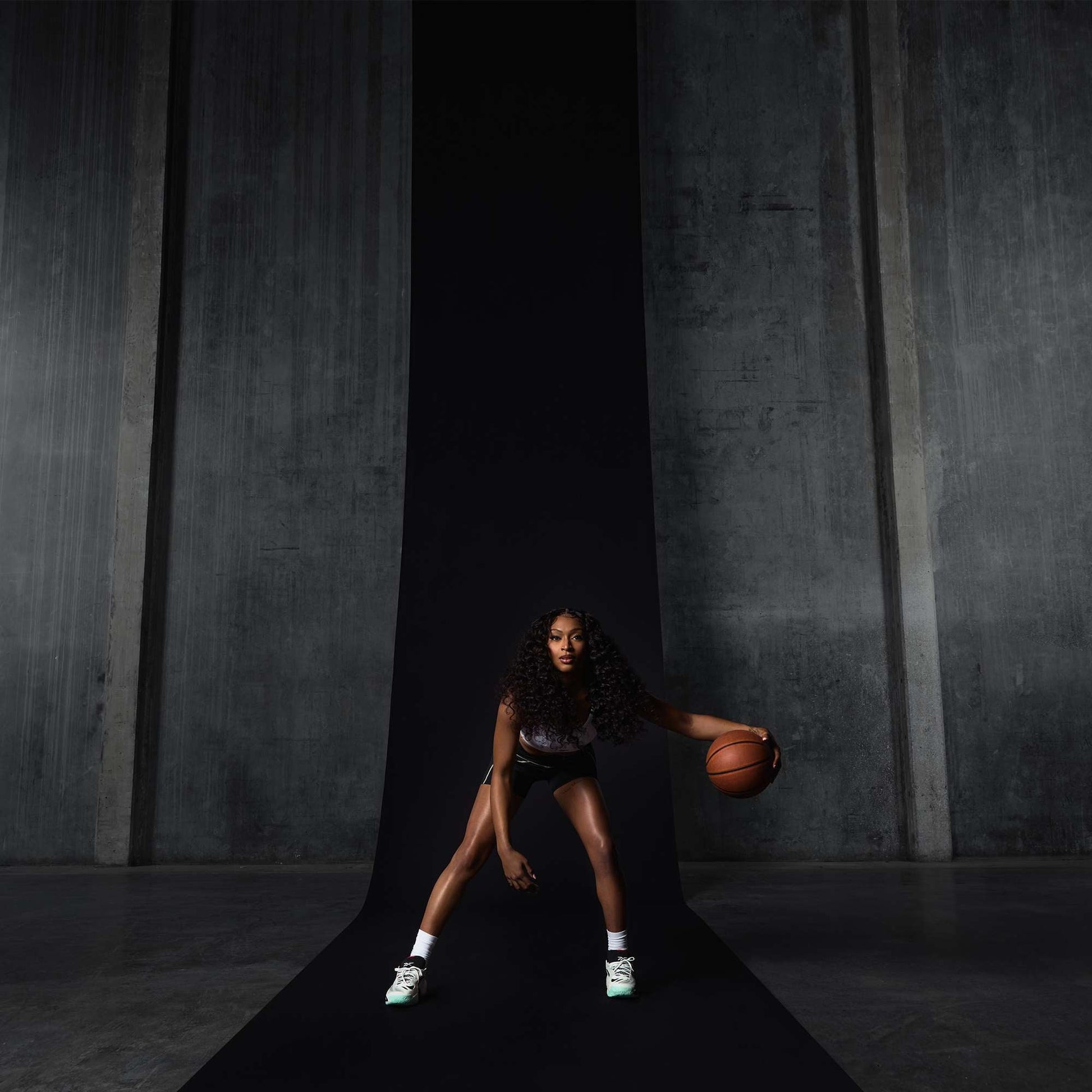 A woman with long curly hair dribbles a basketball on a black runway in front of gray concrete walls, wearing a sports outfit and Reebok Engine A 26 Basketball Shoes designed for energy return and stability, appearing focused and athletic.