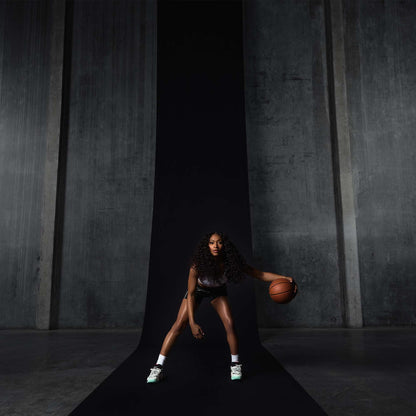 A woman with long curly hair dribbles a basketball on a black runway in front of gray concrete walls, wearing a sports outfit and Reebok Engine A 26 Basketball Shoes designed for energy return and stability, appearing focused and athletic.