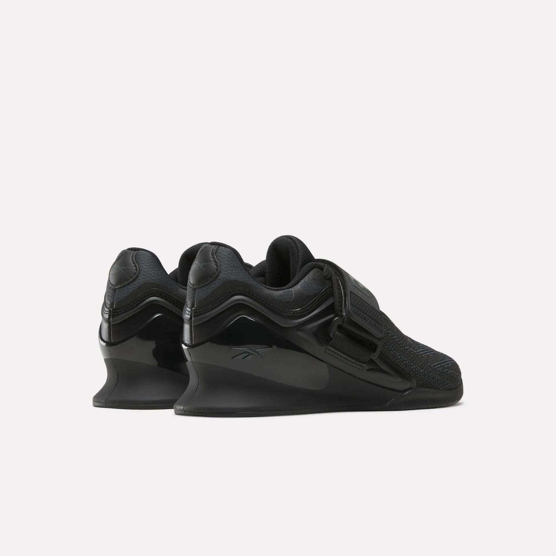 The Reebok Women's Legacy Lifter IV Training Shoes feature elevated heels, mesh uppers, wide straps, and glossy accents, shown from the back and side on a plain white background for support and stability.