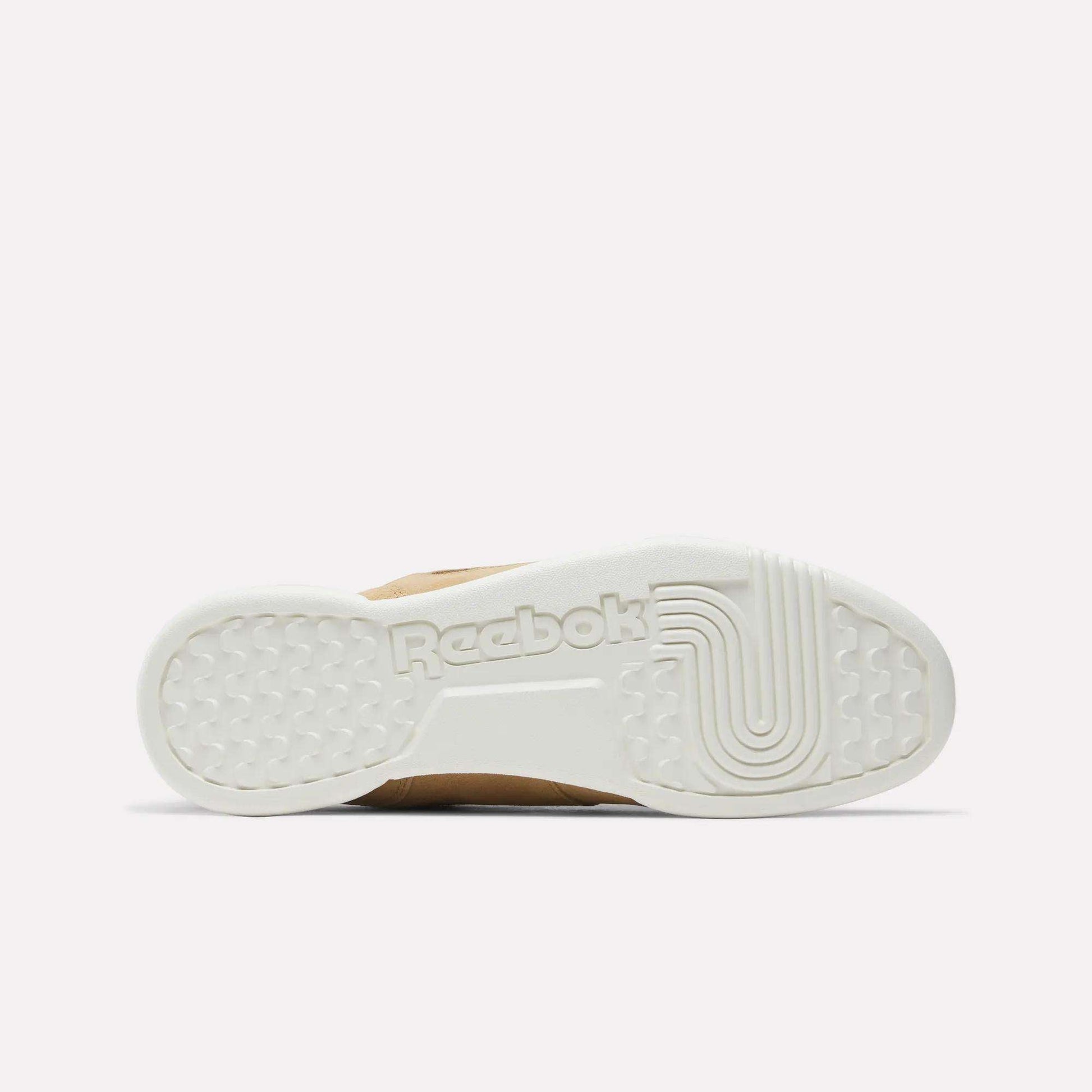 The image shows the white rubber sole of Reebok Workout Plus Shoes from the bottom, featuring textured patterns and an embossed Reebok logo at the center. The plain light pink background evokes classic '80s fitness style.