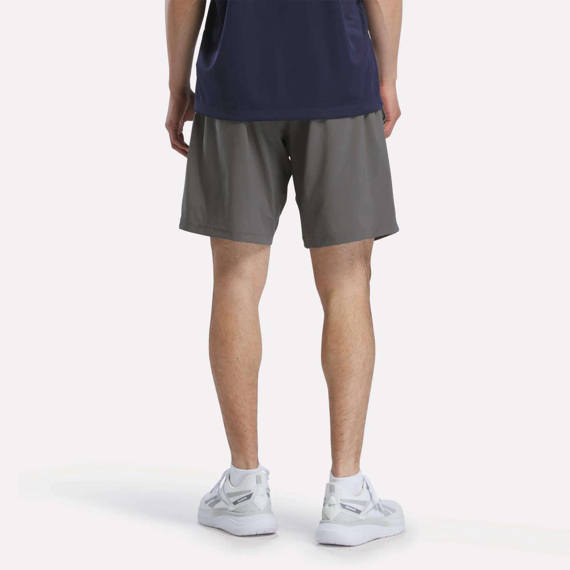 A person faces away, wearing a navy shirt, Reebok Men's Woven Shorts in gray, and white sneakers against a plain background.