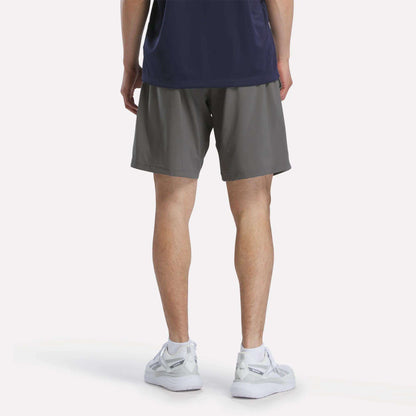 A person faces away, wearing a navy shirt, Reebok Men's Woven Shorts in gray, and white sneakers against a plain background.