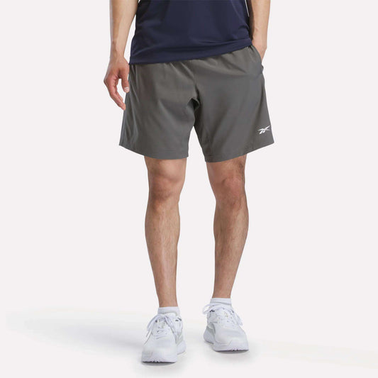 A person wearing a navy shirt, white athletic shoes, and Reebok Men's Woven Shorts in gray with a small white logo stands against a plain white background.