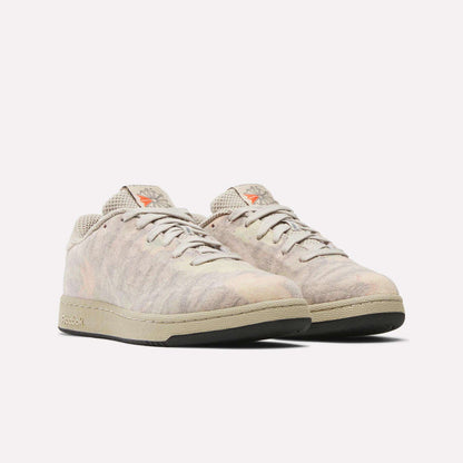 A pair of Reebok x MARKET Club C 85 shoes in light beige with subtle marbled patterns, beige laces, and off-white soles. Court-inspired, they feature the Reebok logo and an orange star emblem on the tongue and side.