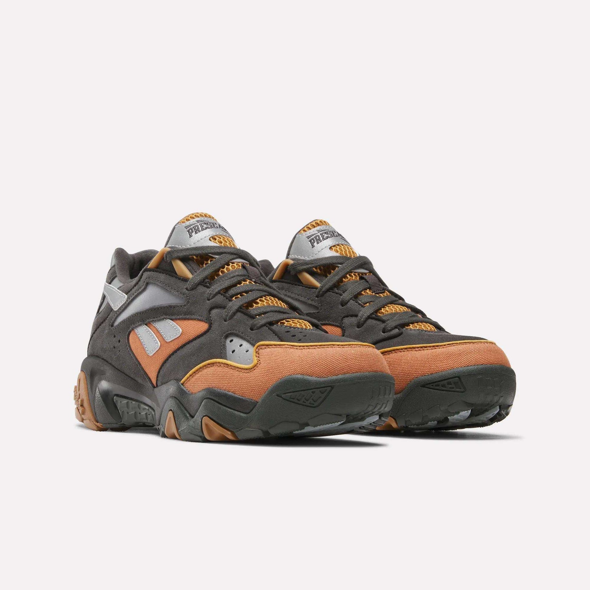 The Reebok x MARKET Preseason 94 Low Shoes feature dynamic black, brown, and gray color blocking with orange accents and chunky soles, showcased against a plain white background.