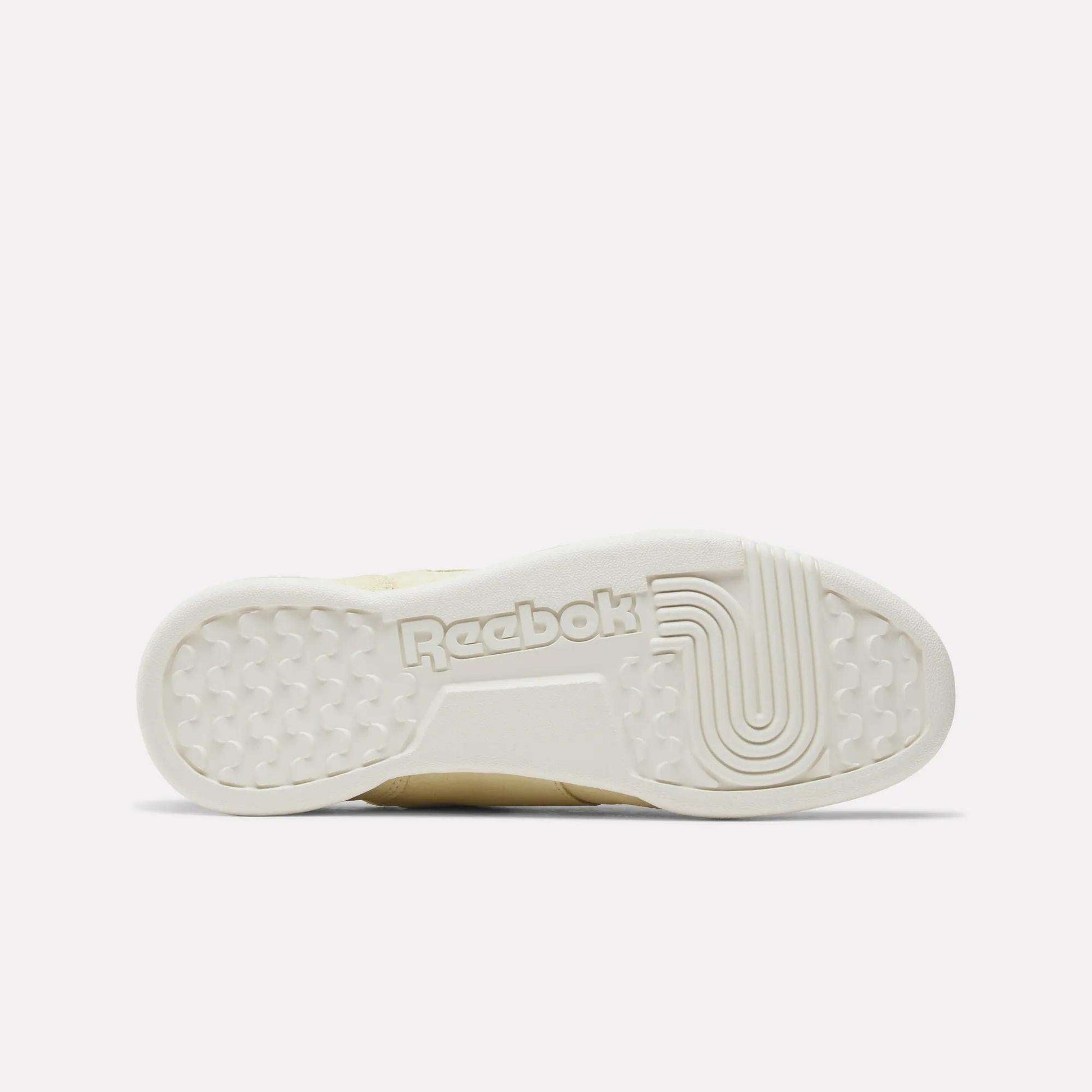 The image displays the outsole of the Reebok Women's Workout Plus Shoes in off-white, highlighted by textured grip patterns and a prominent Reebok logo at the center, reflecting classic '80s fitness style.