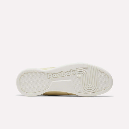 The image displays the outsole of the Reebok Women's Workout Plus Shoes in off-white, highlighted by textured grip patterns and a prominent Reebok logo at the center, reflecting classic '80s fitness style.