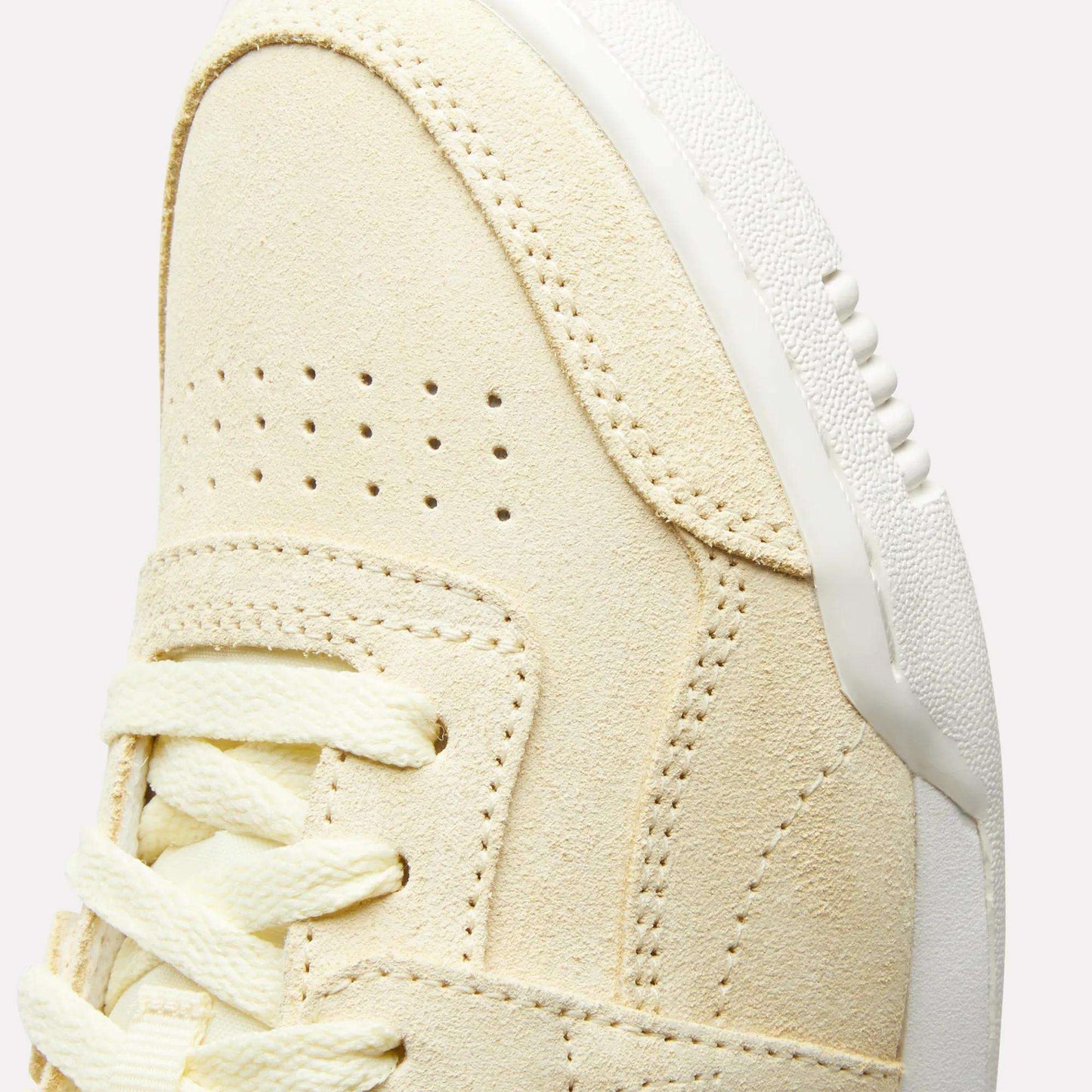 Close-up of the upper front of the Reebok Women's Workout Plus Shoes, featuring beige suede, double perforations, cream laces, and a white textured rubber sole—a classic '80s-inspired fitness style.