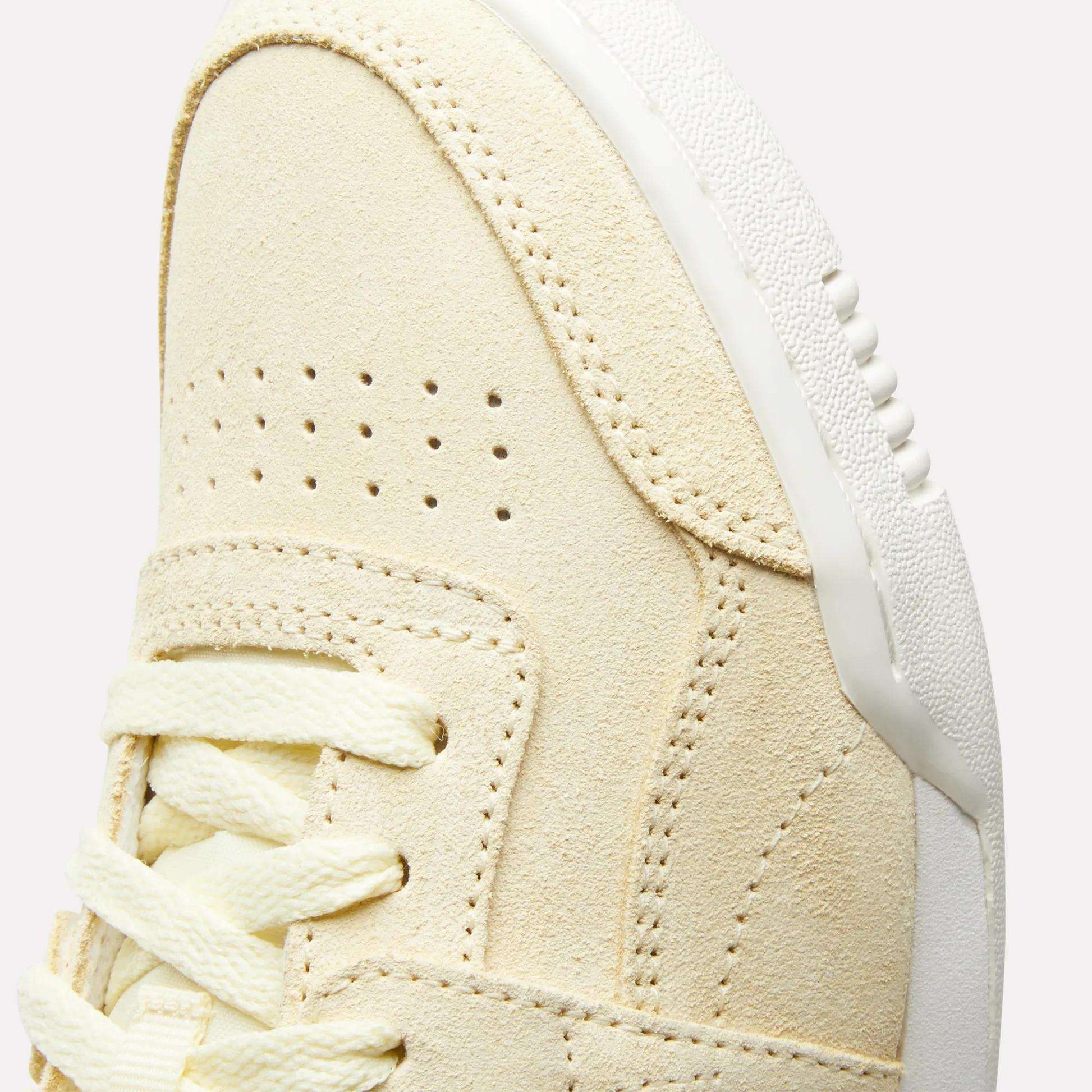 Close-up of the upper front of the Reebok Women's Workout Plus Shoes, featuring beige suede, double perforations, cream laces, and a white textured rubber sole—a classic '80s-inspired fitness style.