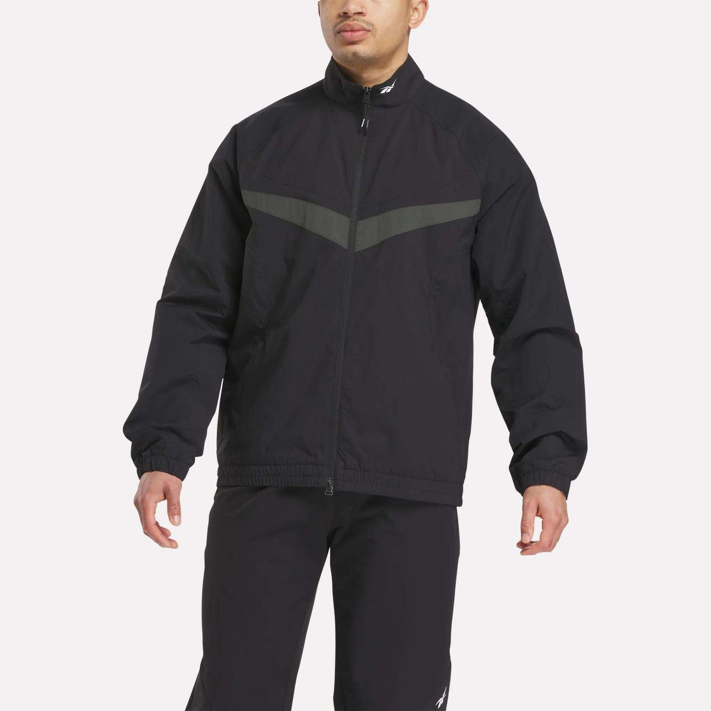 A person wearing the Reebok Warm Up Track Jacket in black with a gray chest stripe and matching black pants, standing against a plain light background.