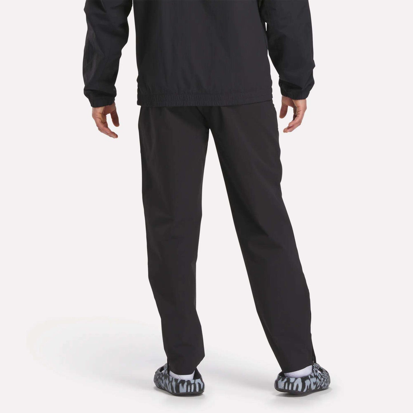 A person stands facing away in Reebok Track Pants, paired with a black jacket and black-and-white patterned slip-on shoes, against a plain light background.
