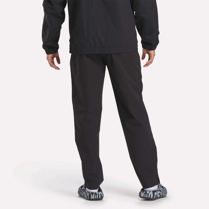A person stands facing away in Reebok Track Pants, paired with a black jacket and black-and-white patterned slip-on shoes, against a plain light background.
