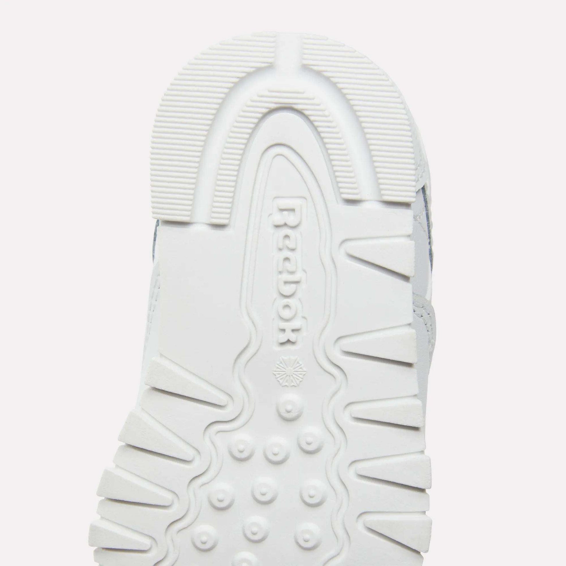 Close-up of the white rubber outsole on Reebok Kids' Classic Leather Shoes, showing the Reebok logo and a textured tread with ridges, dots, and geometric patterns for grip—ideal for everyday wear.