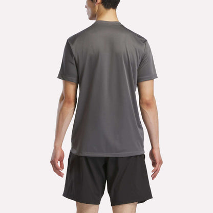 A person stands with their back to the camera, wearing a Reebok Men's Short Sleeve Tech Training T-Shirt in gray and black athletic shorts, highlighting its sweat-wicking comfort for active workouts against a plain light background.