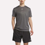 A person wears a Reebok Men's Short Sleeve Tech Training T-Shirt, featuring sweat-wicking fabric, and black shorts with small white logos, standing against a plain white background.