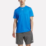 A man stands against a white background wearing a Reebok Men's Short Sleeve Tech Training T-Shirt in bright blue with RBK-DRY fabric and gray shorts, both featuring a small white Reebok logo.