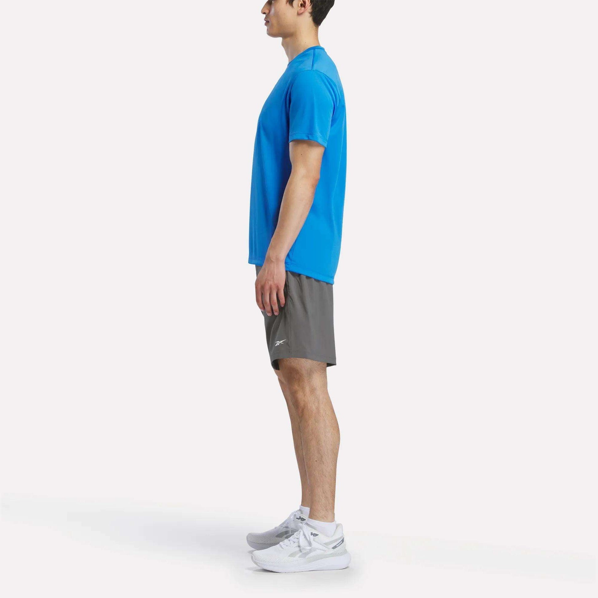A person stands in profile facing left, wearing a blue Reebok Men's Short Sleeve Tech Training T-Shirt, gray shorts, and white athletic shoes against a plain white background.