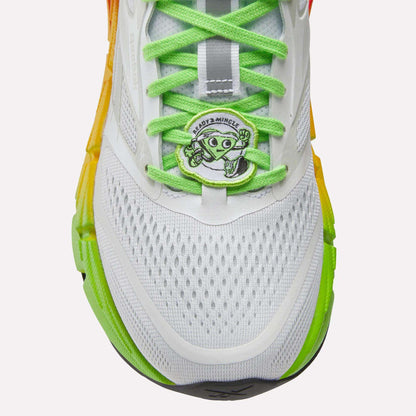 Close-up of Reebok Women's FloatZig 2 SoleMates Running Shoes in white with bright green laces, orange-yellow gradient sides, and a lightweight foam midsole. Features a cartoon alien peace-sign tag reading ANDO2+MINGLE.