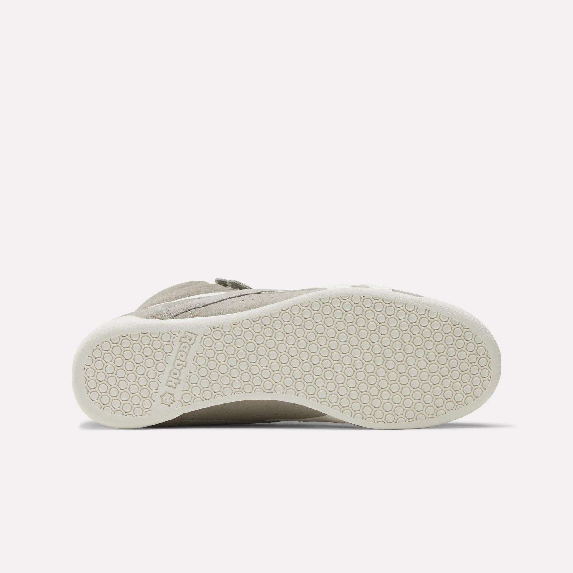 The image shows the bottom view of a beige Women's F/S Hi Shoe by Reebok, with a white sole featuring a hexagonal pattern and the Reebok name embossed near the heel, reflecting popular high top sneaker design trends.