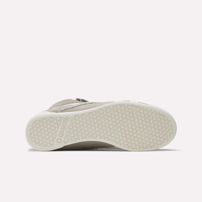 The image shows the bottom view of a beige Women's F/S Hi Shoe by Reebok, with a white sole featuring a hexagonal pattern and the Reebok name embossed near the heel, reflecting popular high top sneaker design trends.