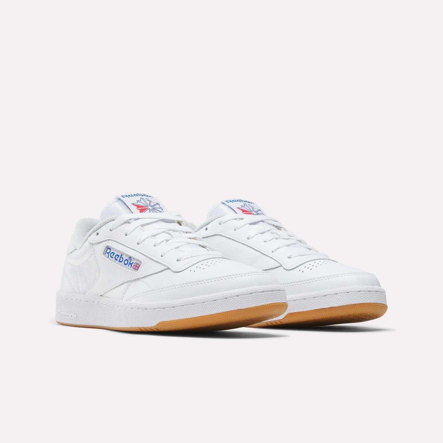 A pair of Reebok Men's Club C 85 Shoes in white leather with gum soles, featuring the Reebok logo and Union Jack on the side and tongue, displayed against a plain light background.