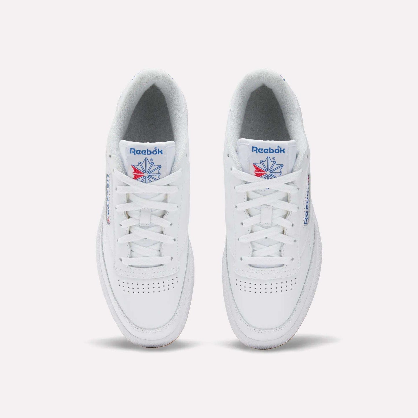 The Reebok Men's Club C 85 Shoes feature a white leather upper, blue Reebok branding, a red and blue star logo on the tongue, white laces, and perforated toe details for a classic tennis-inspired look.