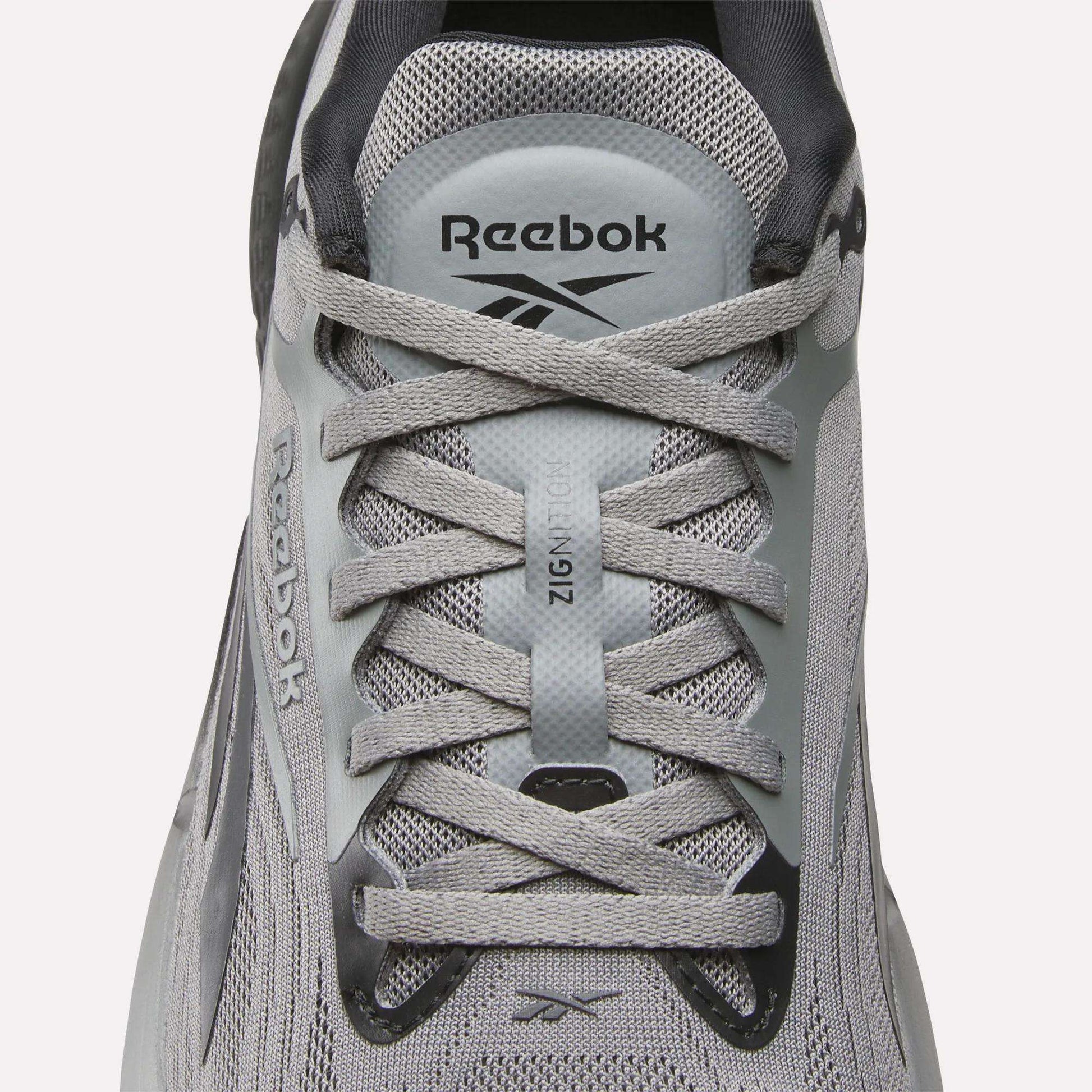 A close-up of Reebok Men's Zignition Running Shoes highlights the laces, mesh upper, and Reebok branding on the tongue and side, designed for cushioned comfort.