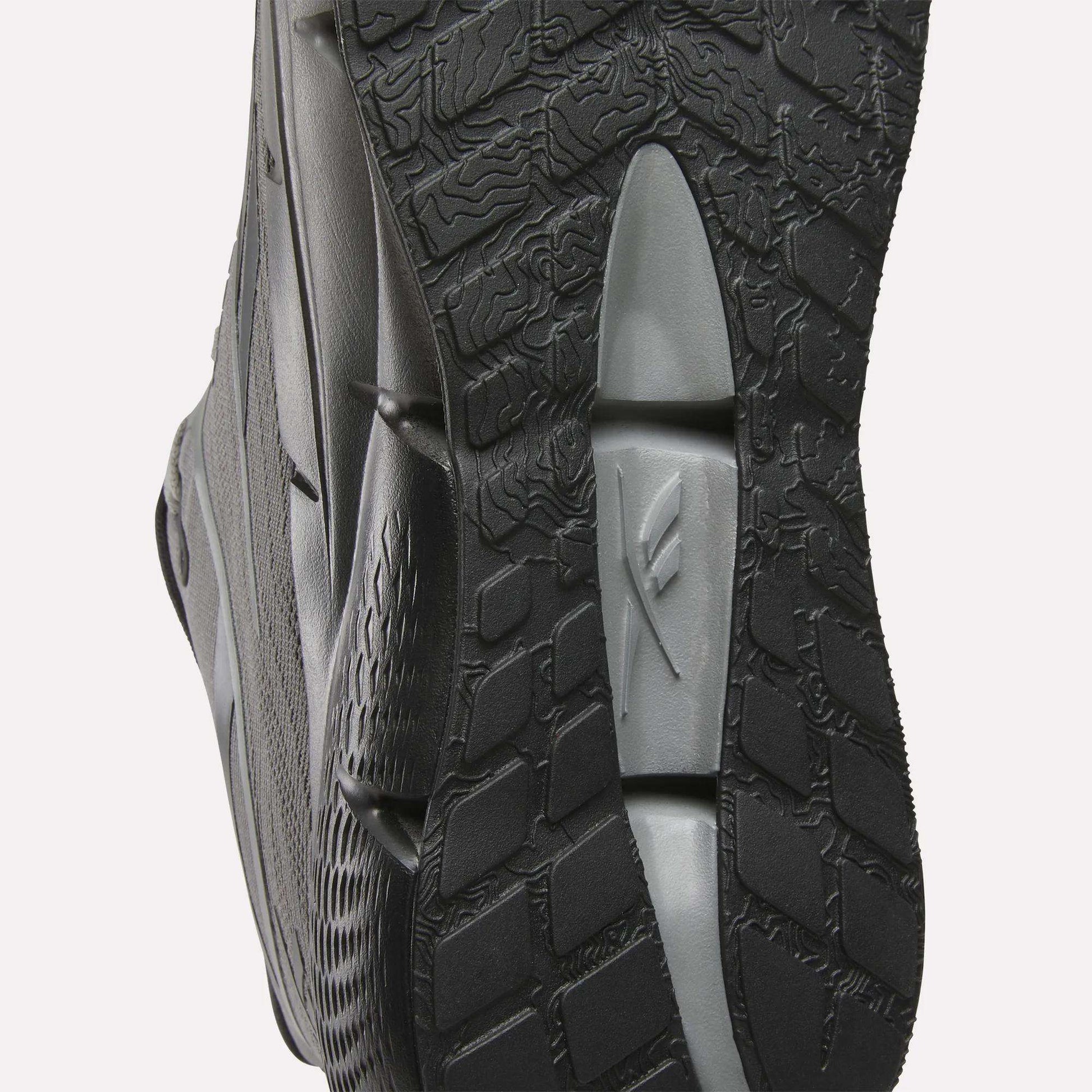 Close-up of the sole of Reebok Men's Zignition Running Shoes in black and gray, highlighting textured patterns and grooves for grip. Wavy and grid-like designs offer optimal support in these cushioned running shoes.