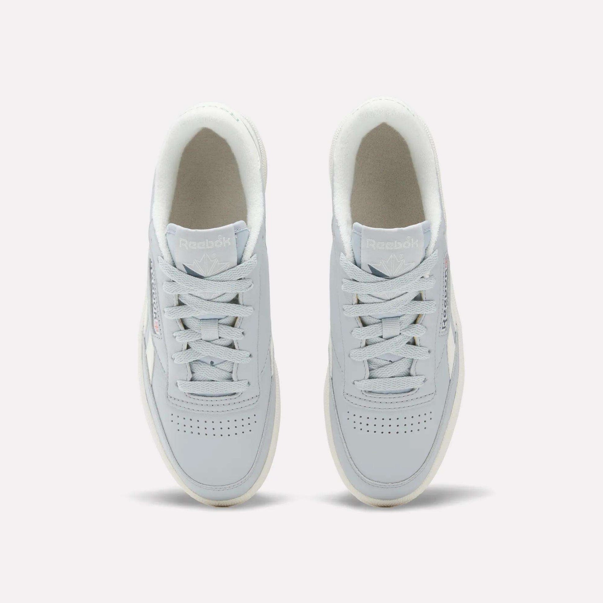 Top view of a pair of Reebok Women's Club C Revenge Shoes in light gray, featuring white soles and laces, placed side by side on a plain background. These casual shoes showcase vintage court style for a timeless look.