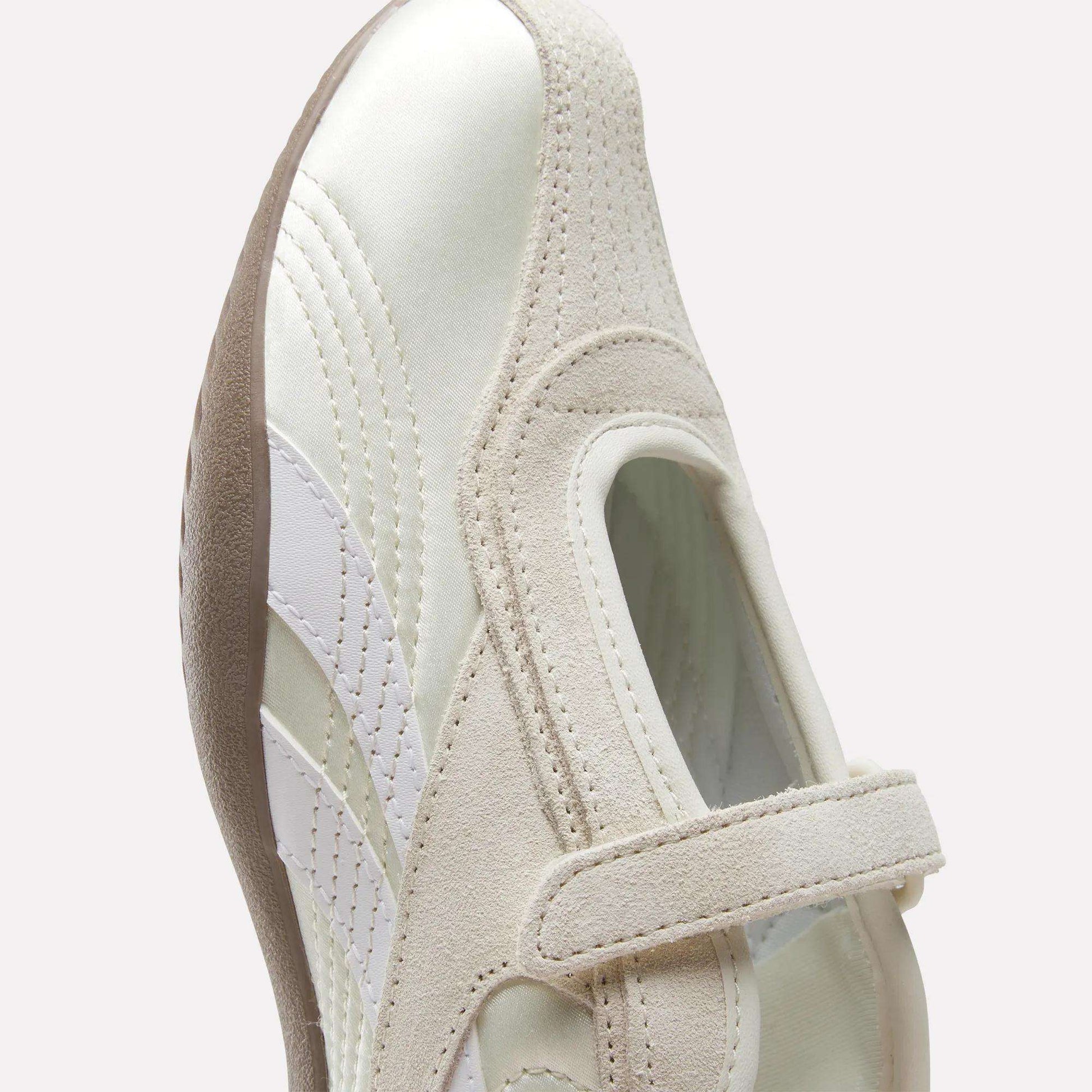 A close-up of the Reebok Women's Ultra Lo Mary Jane Shoes in white and beige, featuring a hook-and-loop strap, suede and leather accents, and a textured brown sole, shown against a light background for a modern, low-profile runner look.