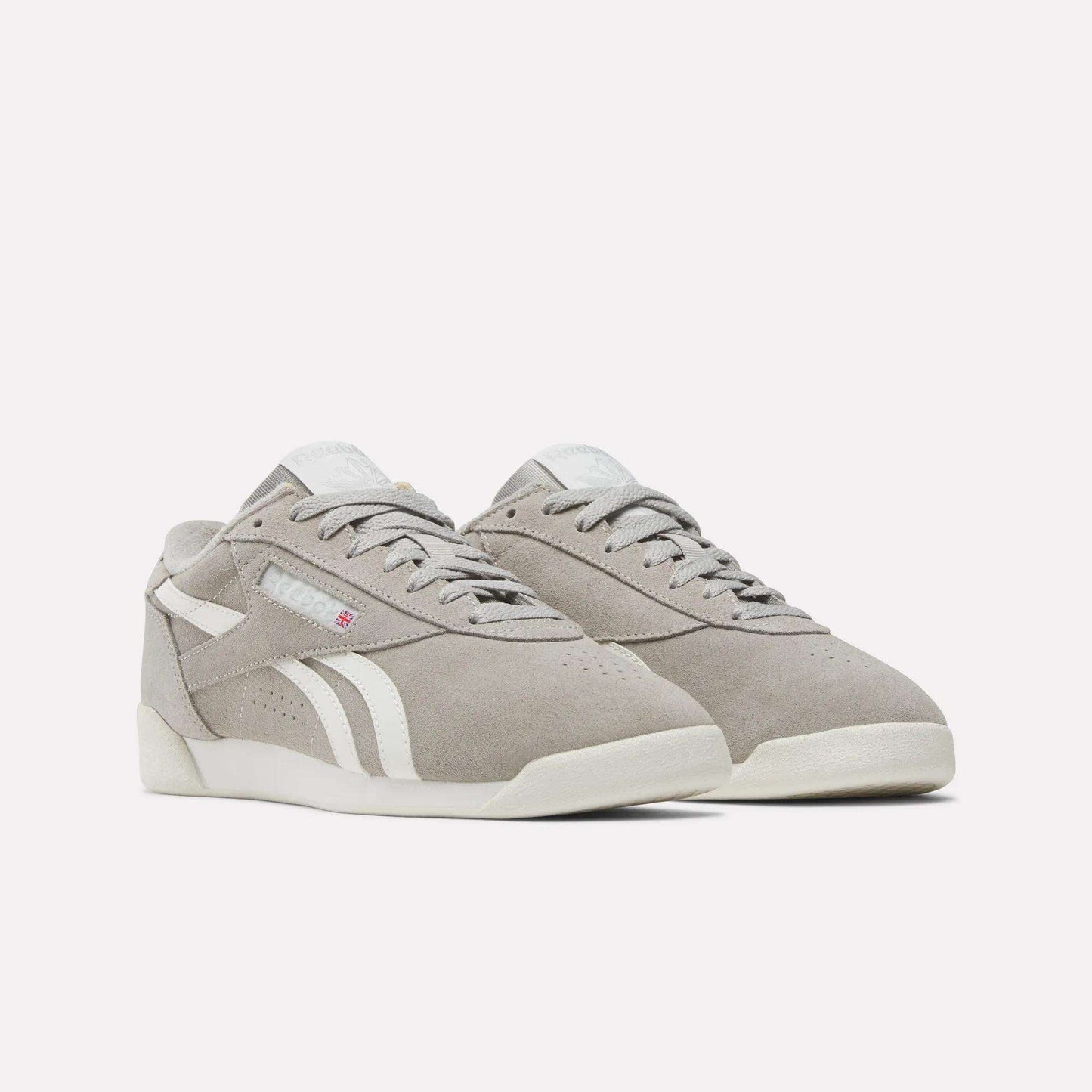 The Reebok Women's Freestyle Lo Shoes are retro sneakers in light gray suede with white soles and side stripes, shown on a simple light background.