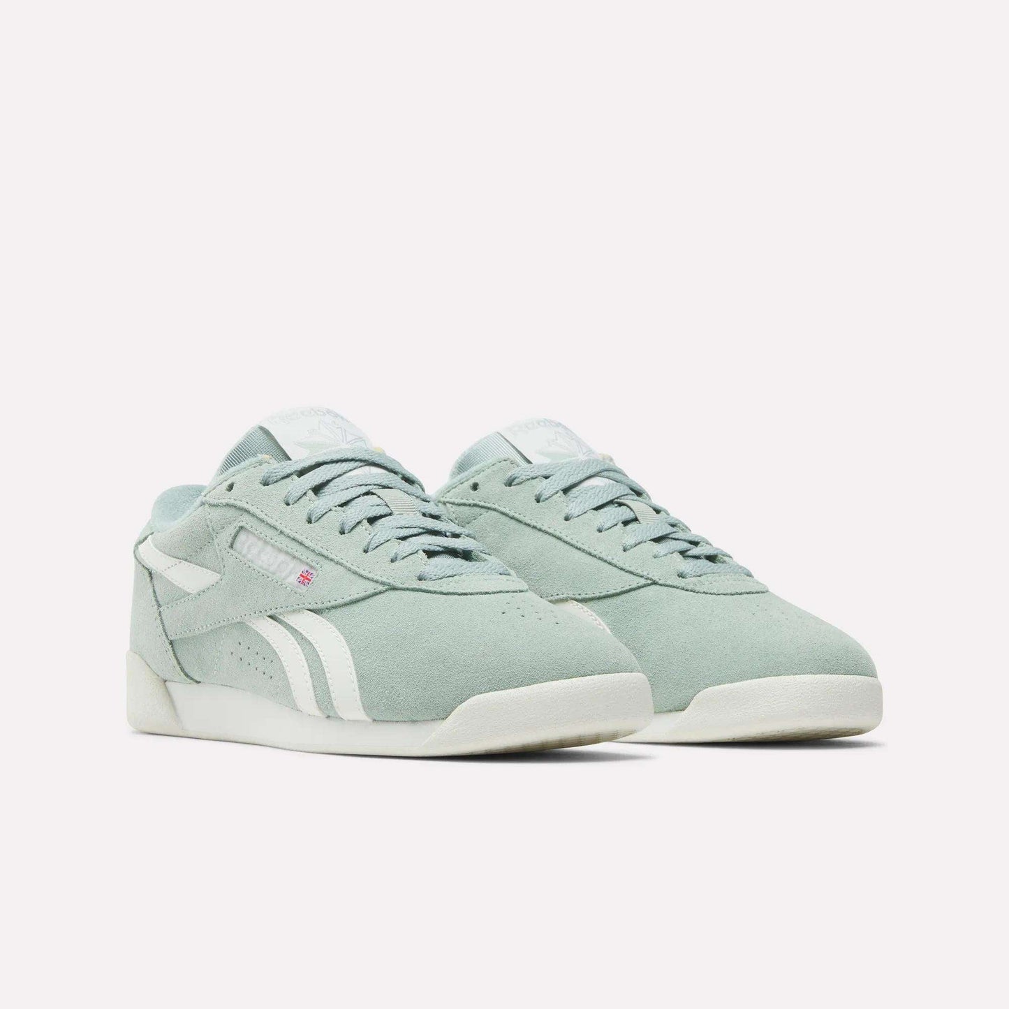 The Reebok Women's Freestyle Lo Shoes in light green suede feature white accents, soles, and side stripes, plus a subtle red logo tag near the laces. The retro sneakers are shown on a plain white background.