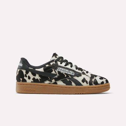 The Reebok Women's Club C Grounds UK Shoes feature a bold black and white cow print upper, black laces, logo accents, and a brown gum sole—making them a standout choice for women’s sneaker collections.