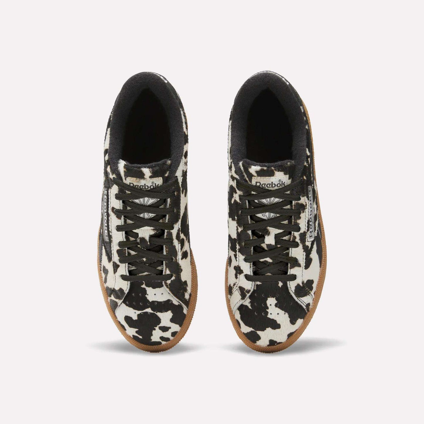 Top view of Reebok Women's Club C Grounds UK Shoes with black and white cow print uppers, black laces, black inner lining, white midsoles, and tan rubber outsoles on a plain white background.