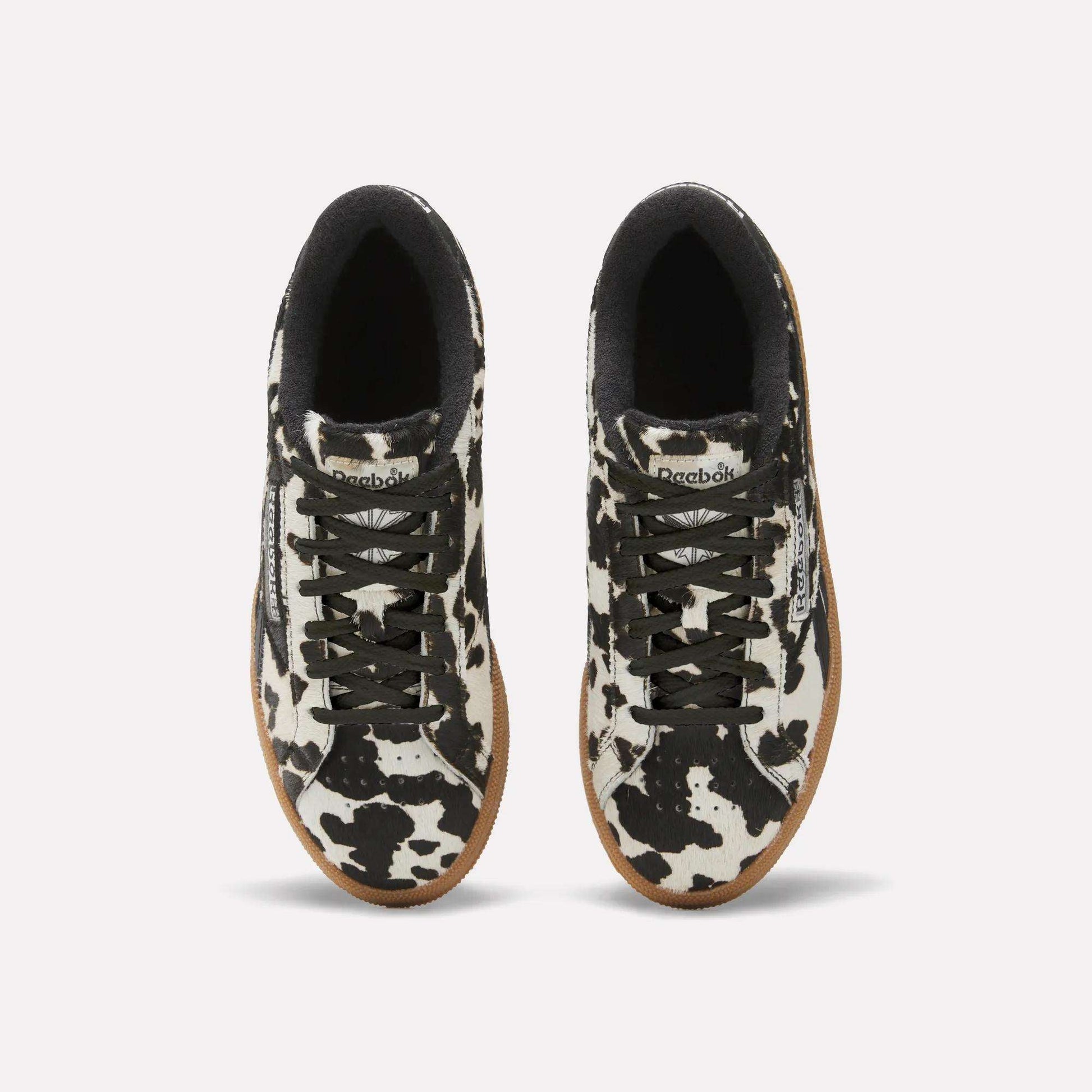Top view of Reebok Women's Club C Grounds UK Shoes with black and white cow print uppers, black laces, black inner lining, white midsoles, and tan rubber outsoles on a plain white background.