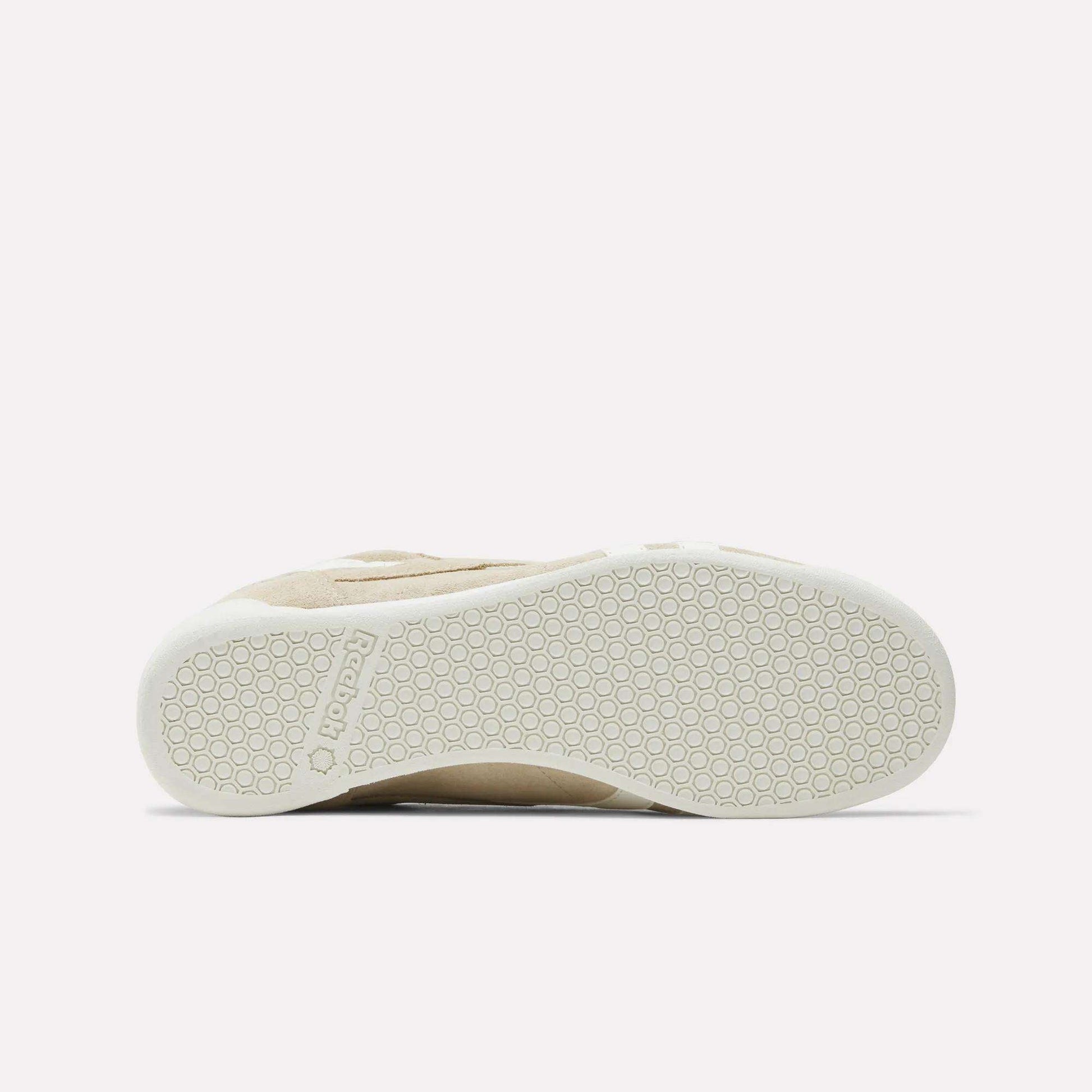 The image shows the bottom view of the Women's Freestyle Lo Shoes by Reebok, featuring a beige suede upper, a white rubber sole with a hexagonal pattern, and "Reebok" embossed near the heel.