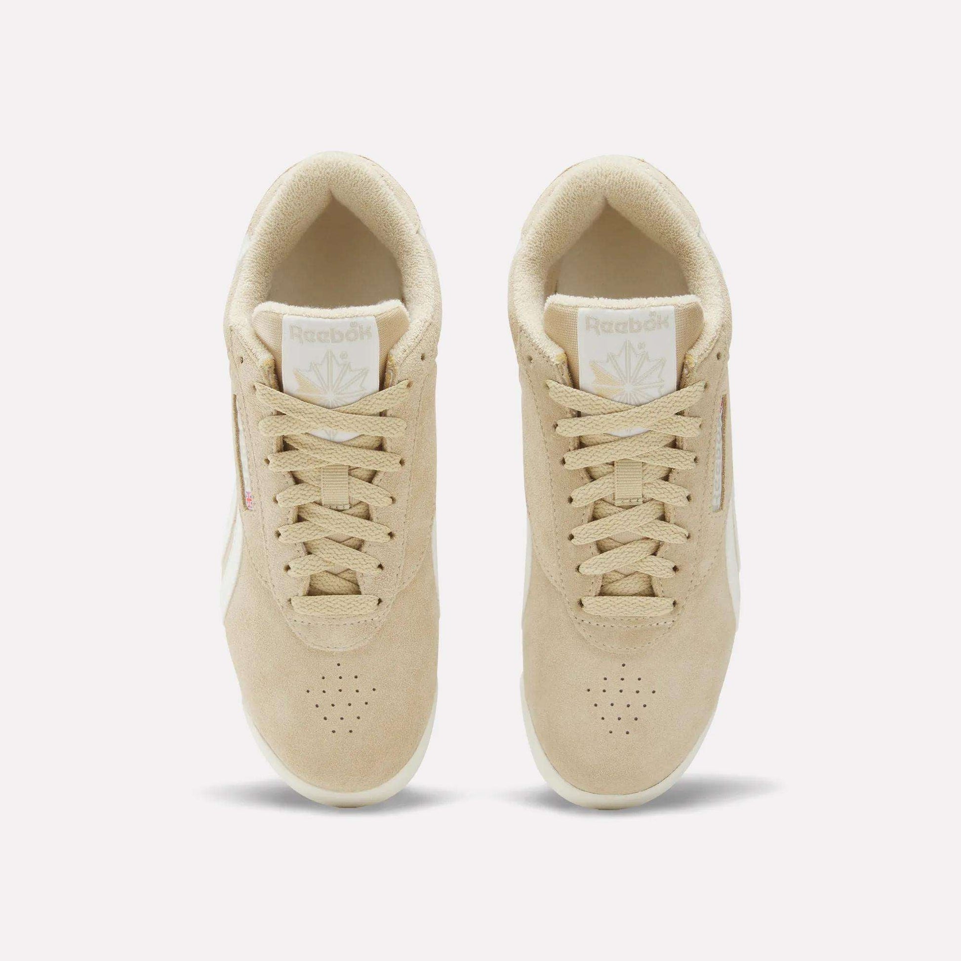 Top view of a pair of Women's Freestyle Lo Shoes by Reebok in beige with white soles and beige laces, featuring the Reebok logo on the tongue and side. The sneakers are placed side by side on a plain white background.