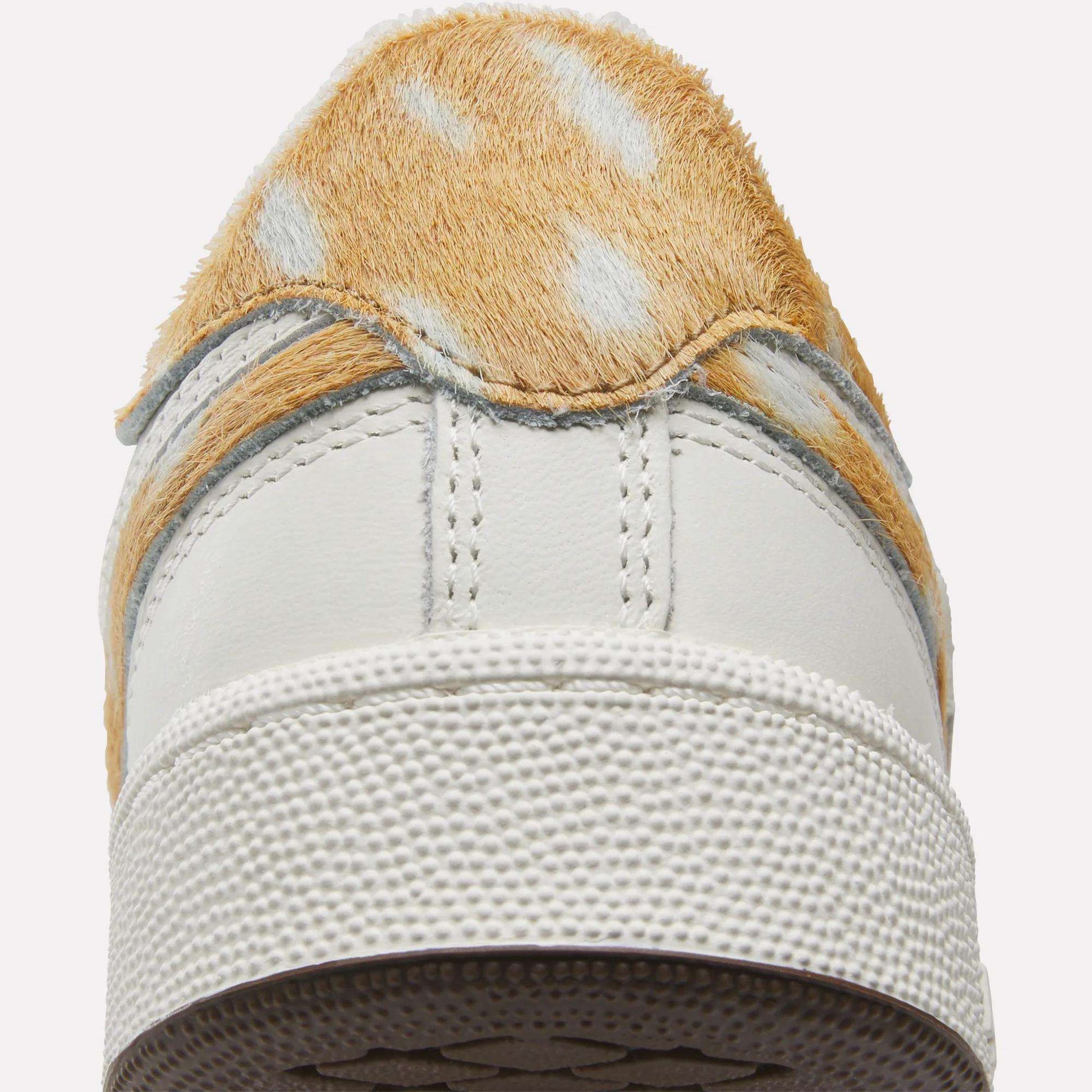 A close-up of the back of the Reebok Club C Revenge Vintage Shoes showcases white sneakers with tan and white faux fur details resembling animal fur, set against a white background. These tennis-inspired shoes feature a leather upper, adding a classic touch to their unique design.