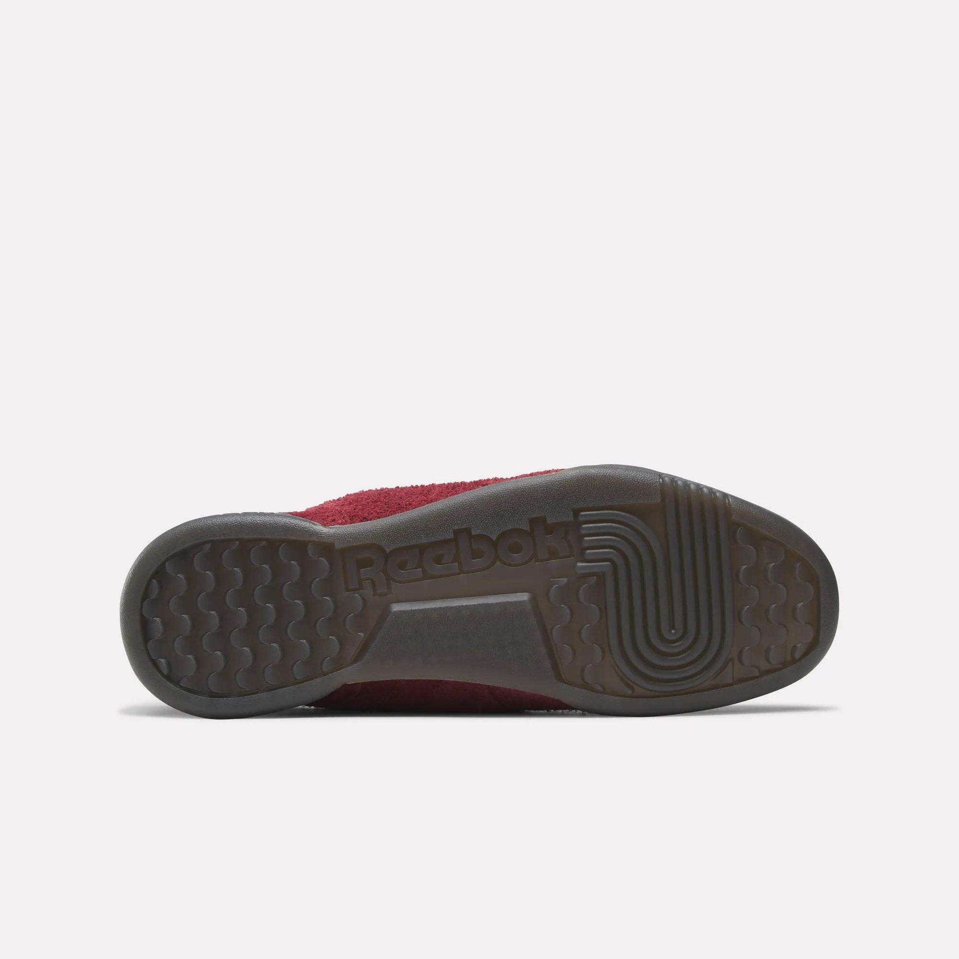 The image shows the bottom view of Reebok Workout Plus Shoes, featuring a dark rubber sole with grip patterns and the Reebok logo. The upper, inspired by ’80s fitness style, is partly burgundy.
