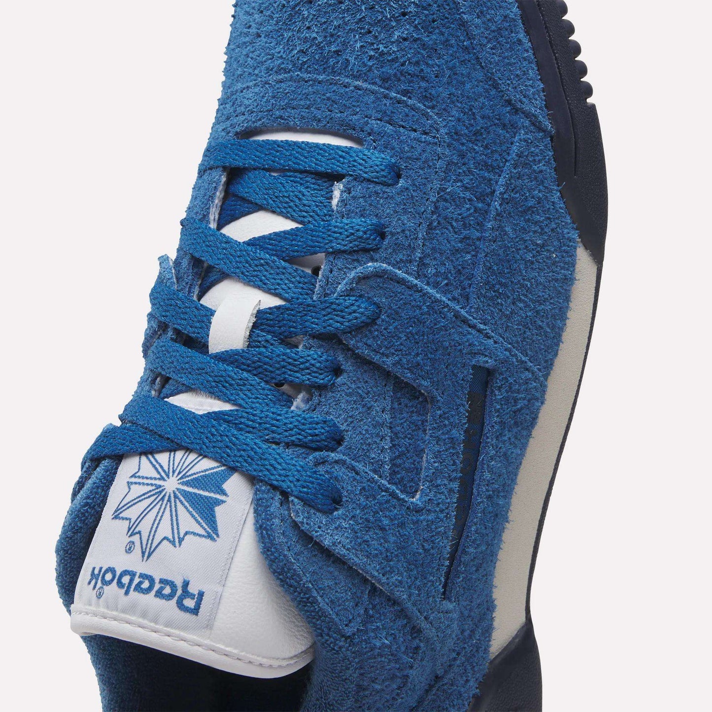 Close-up of Reebok Workout Plus Shoes in blue suede with blue laces and a white tongue featuring the blue Reebok logo and starburst design. This classic '80s-inspired workout sneaker has a white midsole with dark accents.