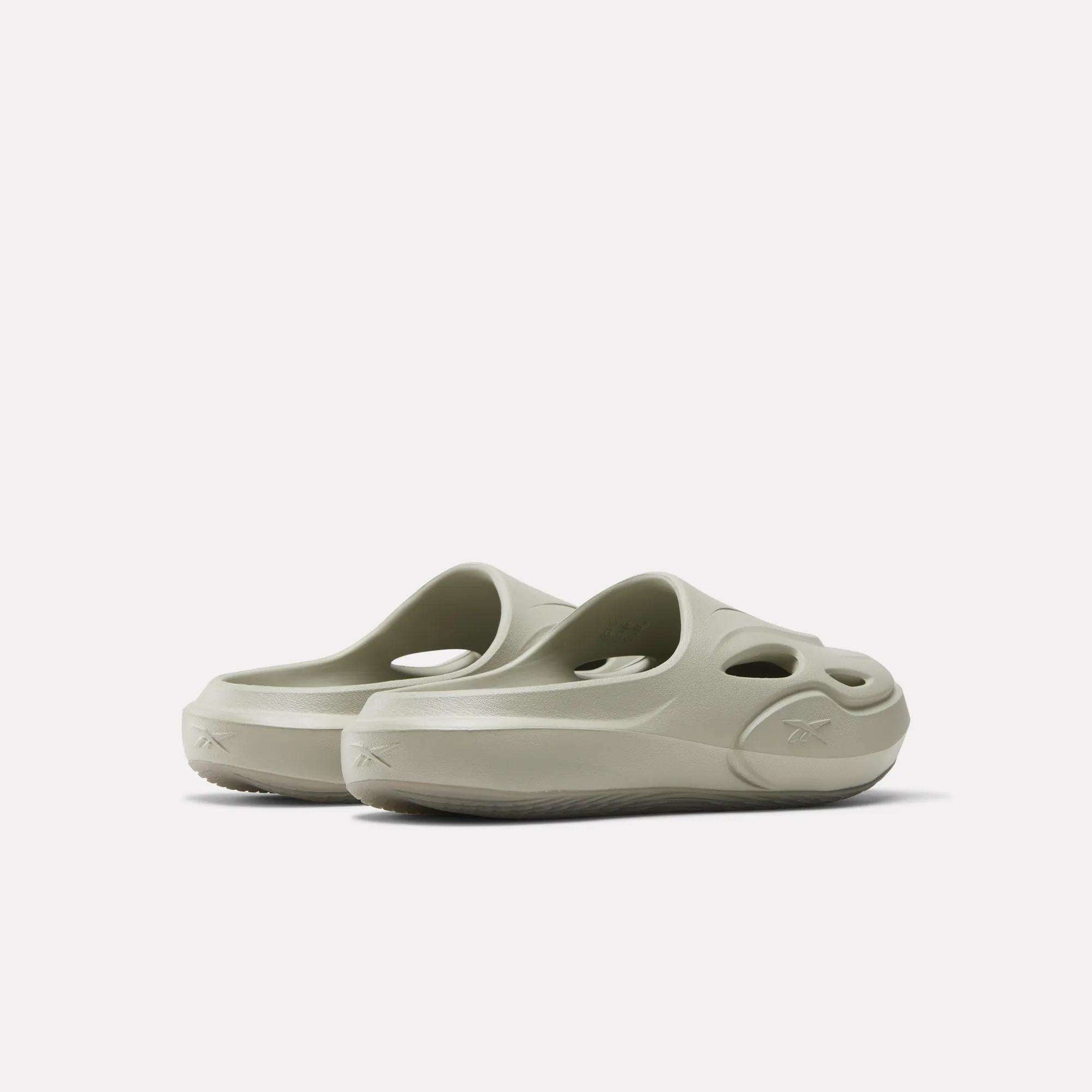 The Reebok DMX Recovery Mules are light beige slip-on EVA shoes with round cut-out holes on the upper and sides, displayed from a rear angle against a plain white background.