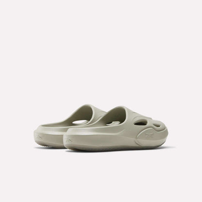 The Reebok DMX Recovery Mules are light beige slip-on EVA shoes with round cut-out holes on the upper and sides, displayed from a rear angle against a plain white background.