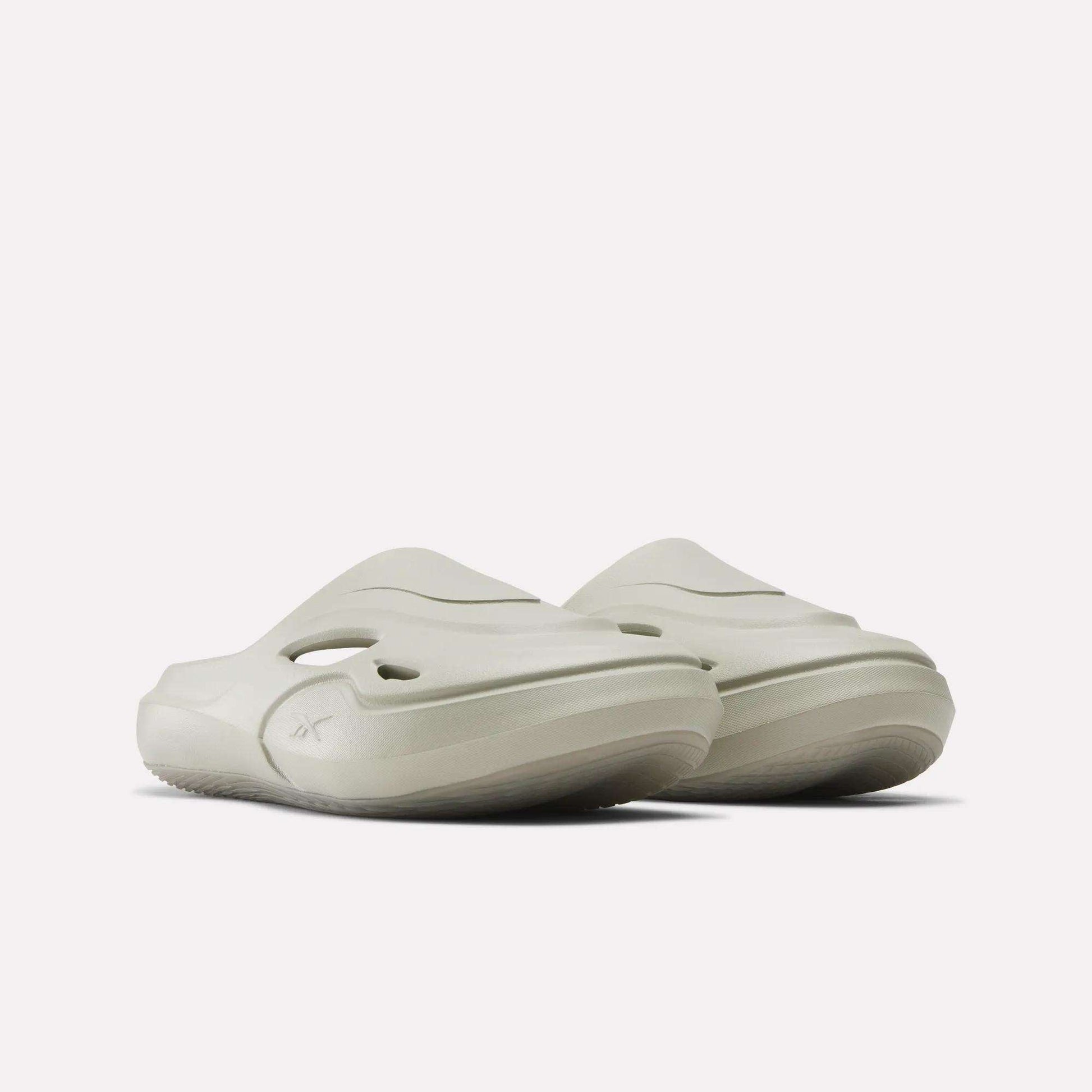 The Reebok DMX Recovery Mules feature a light beige color, chunky molded EVA sole, side cutouts, and a sleek modern design, shown against a plain white background.
