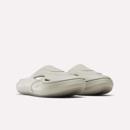 The Reebok DMX Recovery Mules feature a light beige color, chunky molded EVA sole, side cutouts, and a sleek modern design, shown against a plain white background.